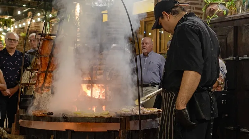Asador trabajando la parrilla circular entre el humo mientras los invitados miran