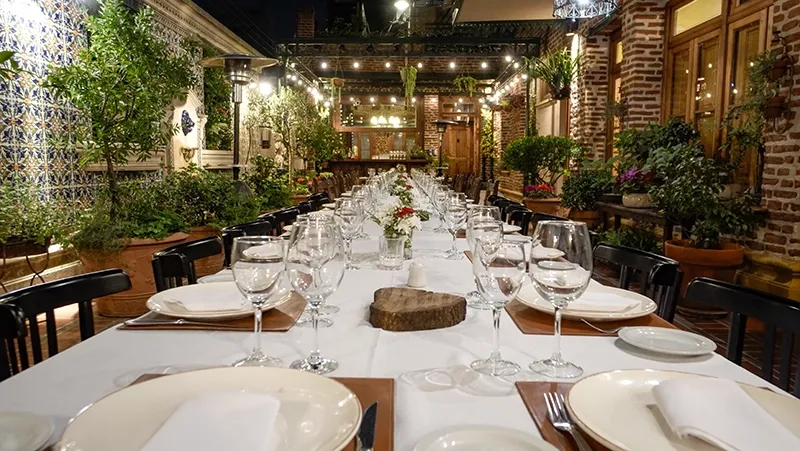 Mesa larga servida en el patio, entre azulejos, plantas y guirnaldas de luces